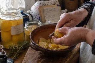Elderly hands slicing preserved lemons over an earthenware bowl in a warmly sunlit, homey kitchen, surrounded by jars and kitchen tools, evoking tradition and quiet concentration.